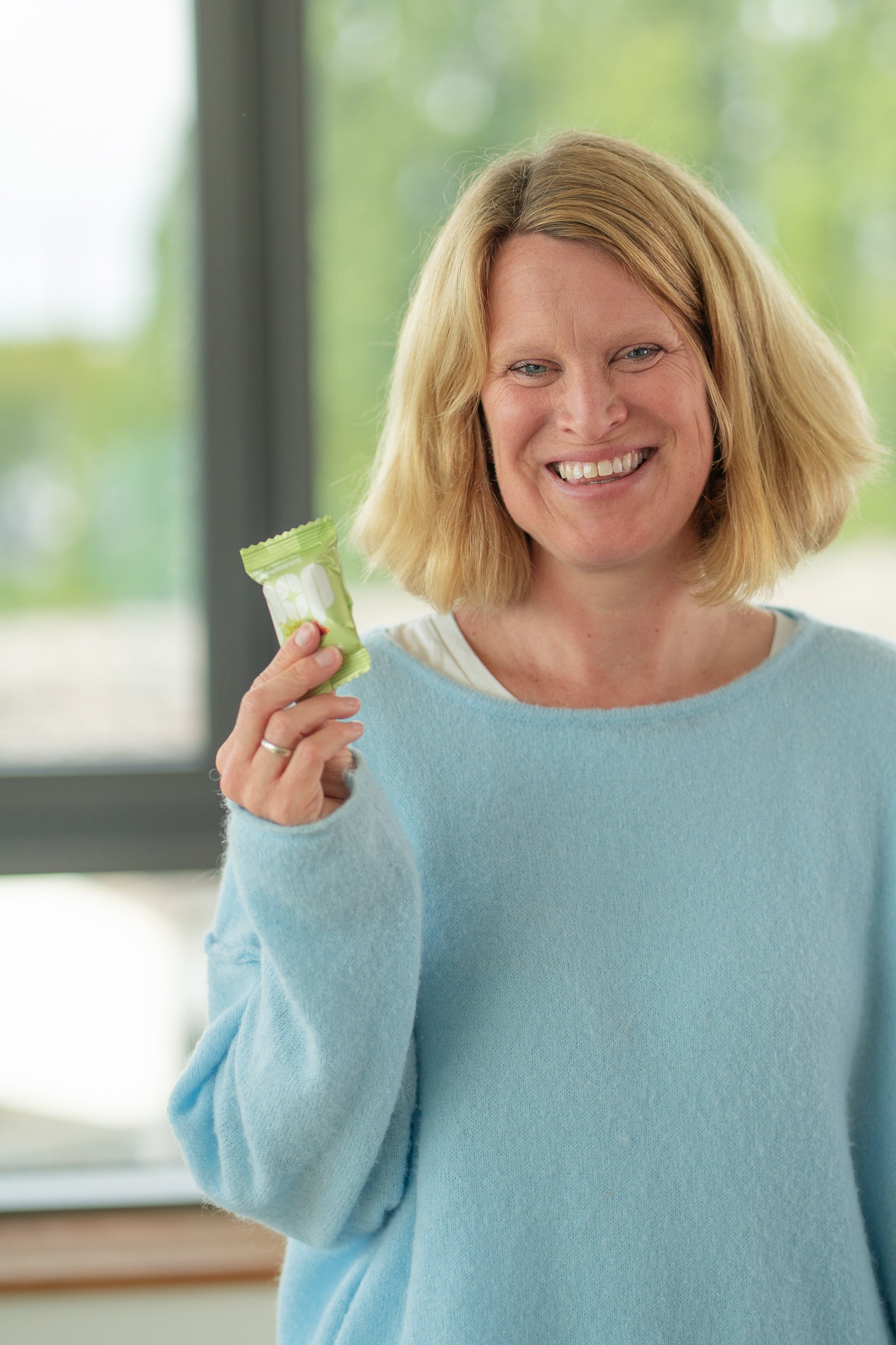 Porträt von Evelyn aus dem POP-Team mit Matcha Mandel Energy Ball in der Hand vor einem Fenster.
